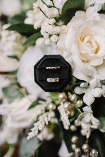 A wedding ring set in a black box placed against a backdrop of white flowers.