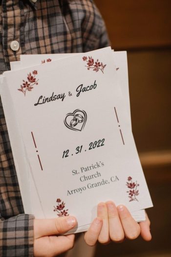 A person holding a wedding program for lindsay and jacob's ceremony on december 31, 2022, at st. patrick's church in arroyo grande, ca.