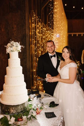 A couple poses for a photograph beside their wedding cake in a decorated venue.