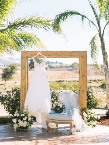 A wedding dress on display under a wooden arch with floral decorations, set against a pastoral landscape.