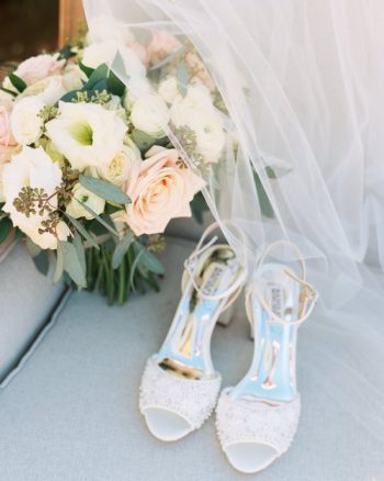A pair of embellished bridal shoes with a bouquet and a veil on a soft blue surface.