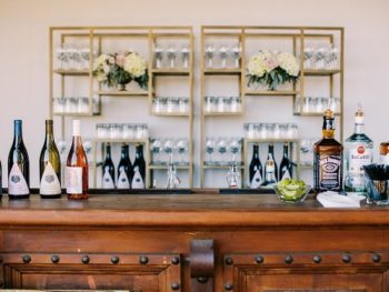 A neatly organized bar with assorted bottles of wine and spirits, glassware on shelves, and sliced limes on the countertop.