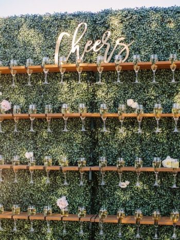 A wall-mounted champagne glass holder with a foliage backdrop and a neon sign reading "cheers.