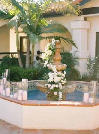 An ornate fountain adorned with white flowers and surrounded by candles in a courtyard setting.