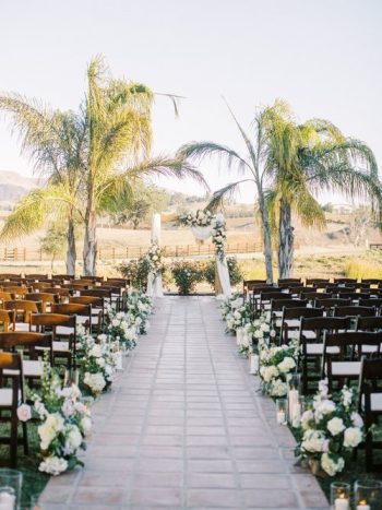 Outdoor wedding aisle lined with chairs, floral arrangements, and lit candles, flanked by palm trees.