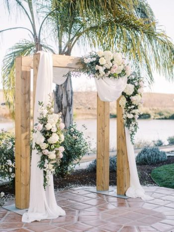 Wooden wedding arch decorated with white flowers and fabric, set outdoors.