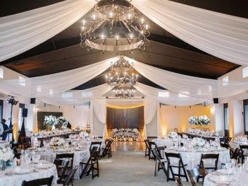 Elegant wedding reception hall decorated with white drapes, chandeliers, and floral centerpieces.