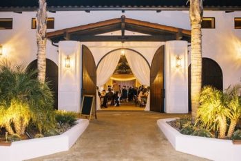 Elegant outdoor evening event under a tent viewed through open doors flanked by palm trees.
