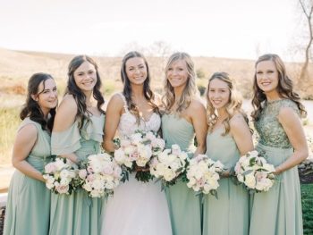 A bride and her bridesmaids smiling for a photo, each holding a bouquet, dressed in pastel dresses for a wedding.
