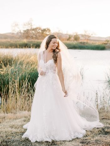 A bride in a white wedding dress with a veil standing outdoors near a body of water.