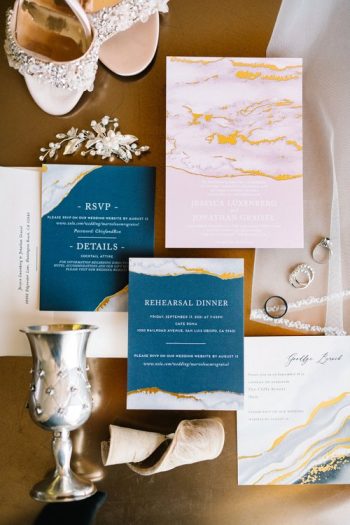 A flat lay of wedding stationery with blue and gold accents, including invitations and rsvp cards, accompanied by bridal accessories such as shoes, a silver goblet, and jewelry.
