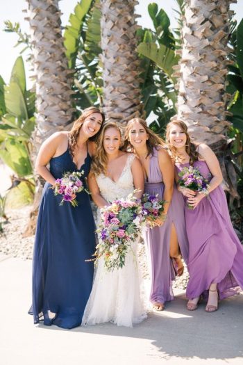 Four women in formal dresses smiling with bouquets, likely at a wedding.