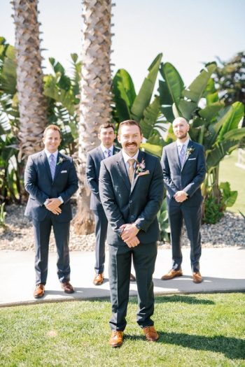 Groom and groomsmen standing outdoors in formal attire with palm trees in the background.