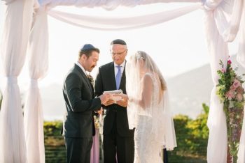 A bride and groom stand under a chuppah with an officiant during a wedding ceremony.