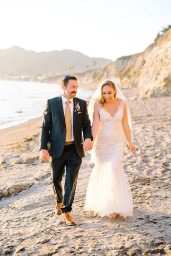 Bride and groom walking hand in hand along a beach.