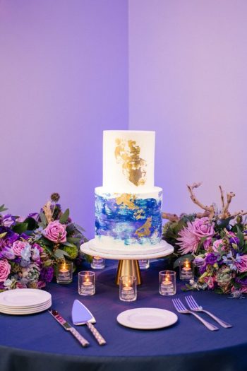 A two-tiered wedding cake with gold leaf accent and blue watercolor design, displayed on a table with floral arrangements and candles.
