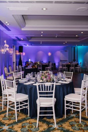 Elegantly set tables with white chairs in a banquet hall with blue lighting.