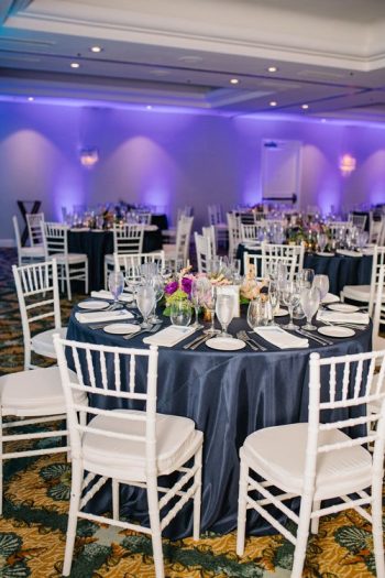 Elegant banquet hall setup with round tables, white chairs, navy tablecloths, and floral centerpieces.