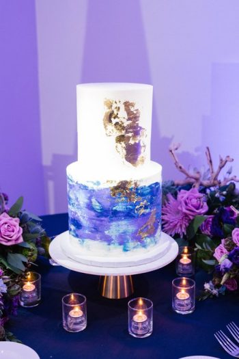 Two-tiered wedding cake with abstract blue and gold design, displayed on a table with lit candles and floral arrangements.