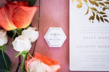 Wedding invitation with a ring in a hexagonal box, accompanied by a rose and petals.