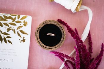 A wedding ring in a circular wooden box placed on a pink surface beside an invitation and a sprig of pink flowers.