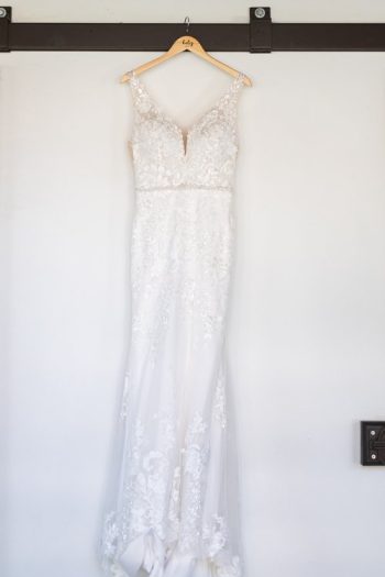 A white bridal gown with lace detailing hanging on a hanger against a plain wall.