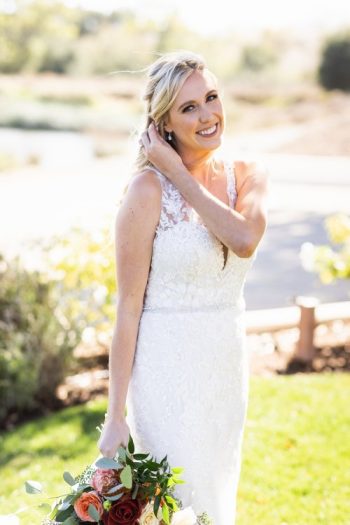 Bride in a lace dress holding a bouquet and smiling outdoors.