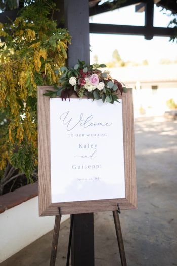 A wedding welcome sign featuring the names 'kaley and guiseppi' with a date, displayed on an easel and adorned with a floral arrangement.