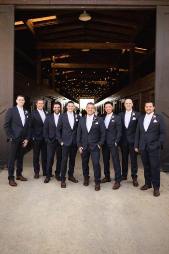 Group of seven men dressed in matching suits and boutonnieres, possibly groomsmen at a wedding, standing confidently in a wooden barn aisle.