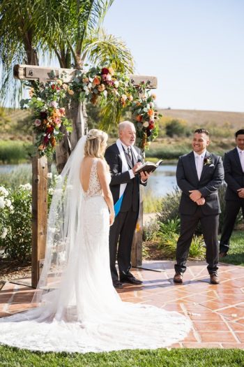 Bride and groom at an outdoor wedding ceremony with an officiant speaking and a guest observing.