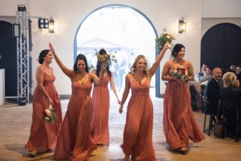Bridesmaids in coral dresses walk down the aisle at a wedding, smiling and carrying bouquets.