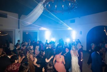 A wedding reception dance floor with guests dancing under blue lights and a chandelier.