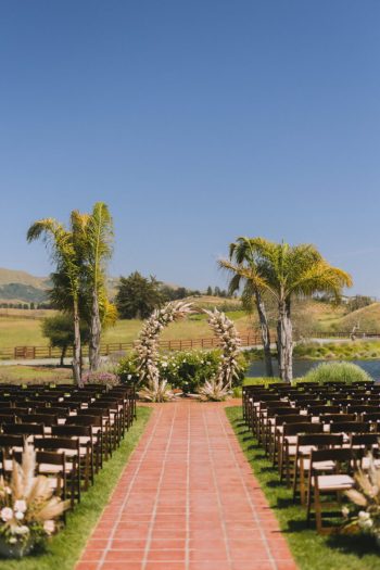 An outdoor wedding aisle with chairs and palm trees leading to an archway, set against a rural landscape.