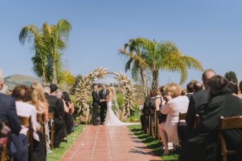 A couple stands at the altar during an outdoor wedding ceremony with guests seated on either side of the aisle.