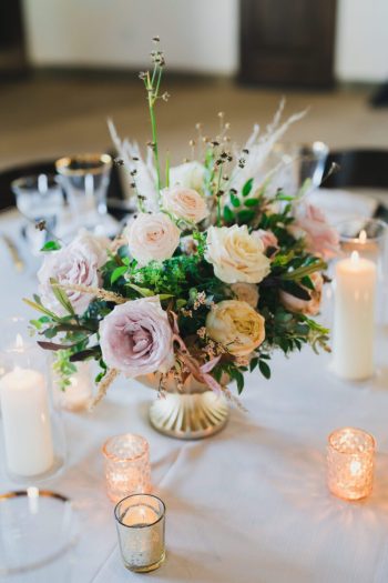 A floral centerpiece with roses and greenery, surrounded by lit candles on a table setting.