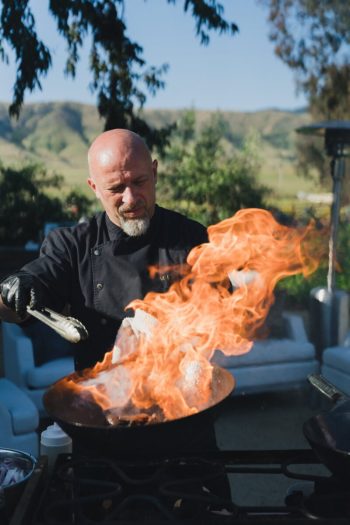 Chef cooking with a large flame in a wok outdoors.