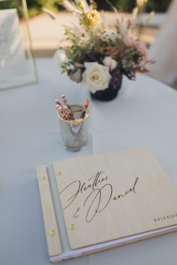 A guest book with the names "heather & daniel" and a date on the cover, placed beside a floral centerpiece and a container of pens.