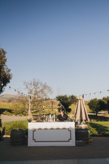 Outdoor event setup with a bartender behind a bar stand, string lights, and scenic backdrop.