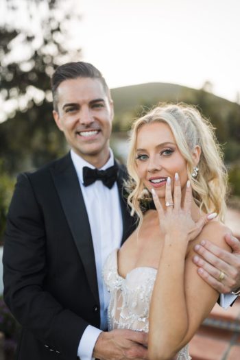 A smiling couple in formal attire with the woman showing off her engagement ring.