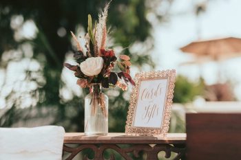 Floral arrangement in a jar beside a framed sign reading "cards & gifts" on a wooden table at an outdoor event.