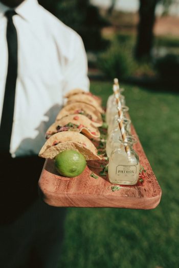 Row of tacos on a wooden serving board with a lime and a bottle of patrón tequila.