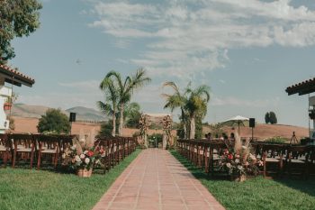 An outdoor wedding ceremony setup with rows of chairs and a floral archway at the end of a red-tiled aisle, surrounded by a serene landscape.