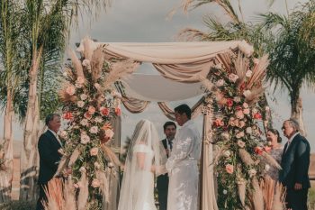 A wedding ceremony outdoors with an officiant, a couple exchanging vows under a floral archway, and guests looking on.