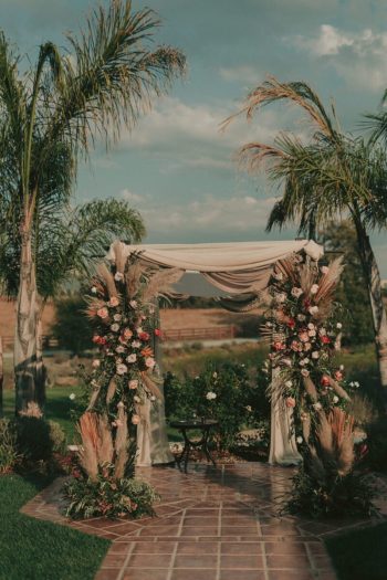 An outdoor wedding altar adorned with floral arrangements and draped fabric, set against a scenic backdrop with palm trees.