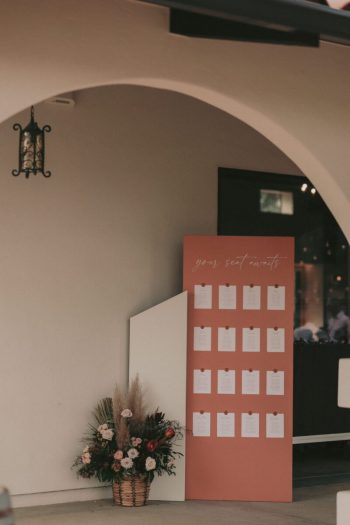 An elegant event setup with a seating chart display next to a floral arrangement at an arched entrance.