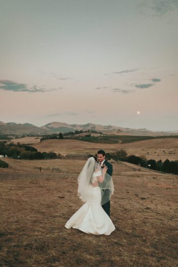 A couple in wedding attire embracing in a rural landscape at dusk.