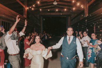 A joyful bride and groom hold hands as they walk through a cheering crowd with string lights above them.